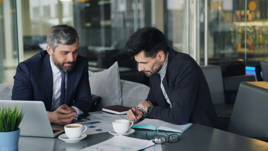 Two businessmen analyze stock market data using a smartphone and laptop in a modern office setting.